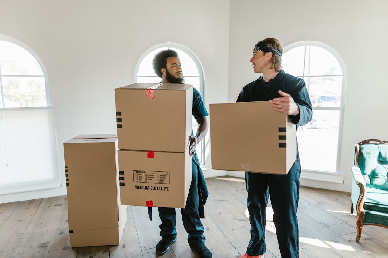 creative-02 Two professional movers carrying cardboard boxes inside a bright room with large windows.